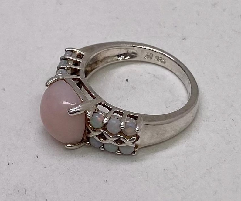 K&ouml;nnte beinhalten: Ein silberner Ring mit einem gro&szlig;en rosafarbenen Opal-Edelstein und kleineren wei&szlig;en Opalen, die in einem filigranen Design eingefasst sind.