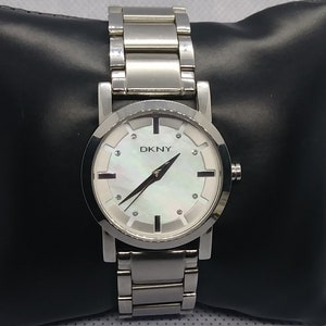 Puede incluir: Un reloj plateado con una esfera blanca y una correa de metal plateado. La esfera del reloj tiene un diseño de nácar con detalles plateados y la palabra "DKNY" impresa en ella.