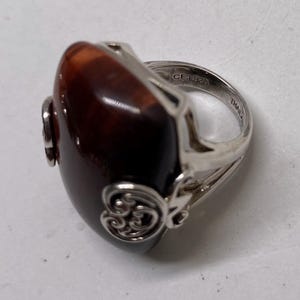May include: A silver ring with a large, oval, polished, brown stone. The ring has decorative silver filigree on the sides and a band marked "CF.925 THAILAND". The stone has a glossy finish.