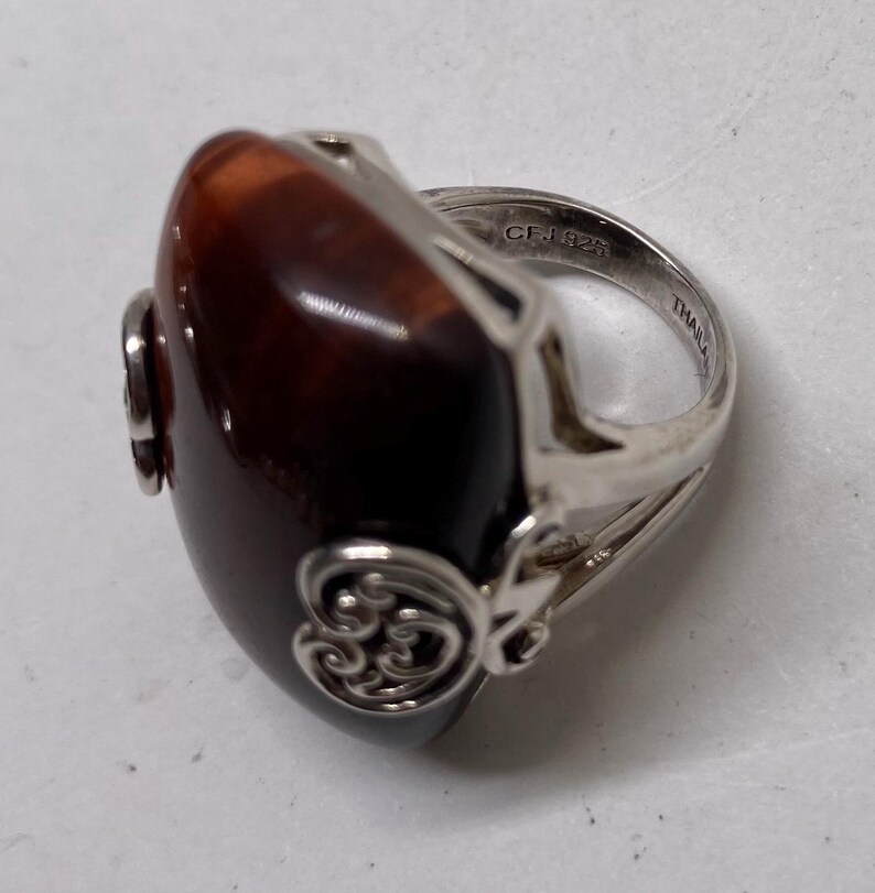 May include: A silver ring with a large, oval, polished, brown stone. The ring has decorative silver filigree on the sides and a band marked "CF.925 THAILAND". The stone has a glossy finish.