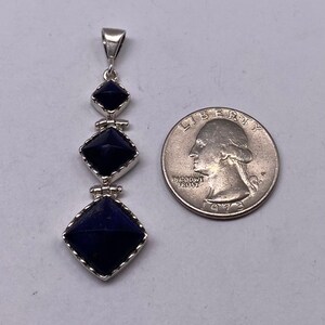 May include: A silver pendant with three dark blue lapis lazuli stones in a square shape, each stone is set in a silver frame with a scalloped edge. The pendant is hanging from a silver bail. The pendant is about the size of a US quarter.