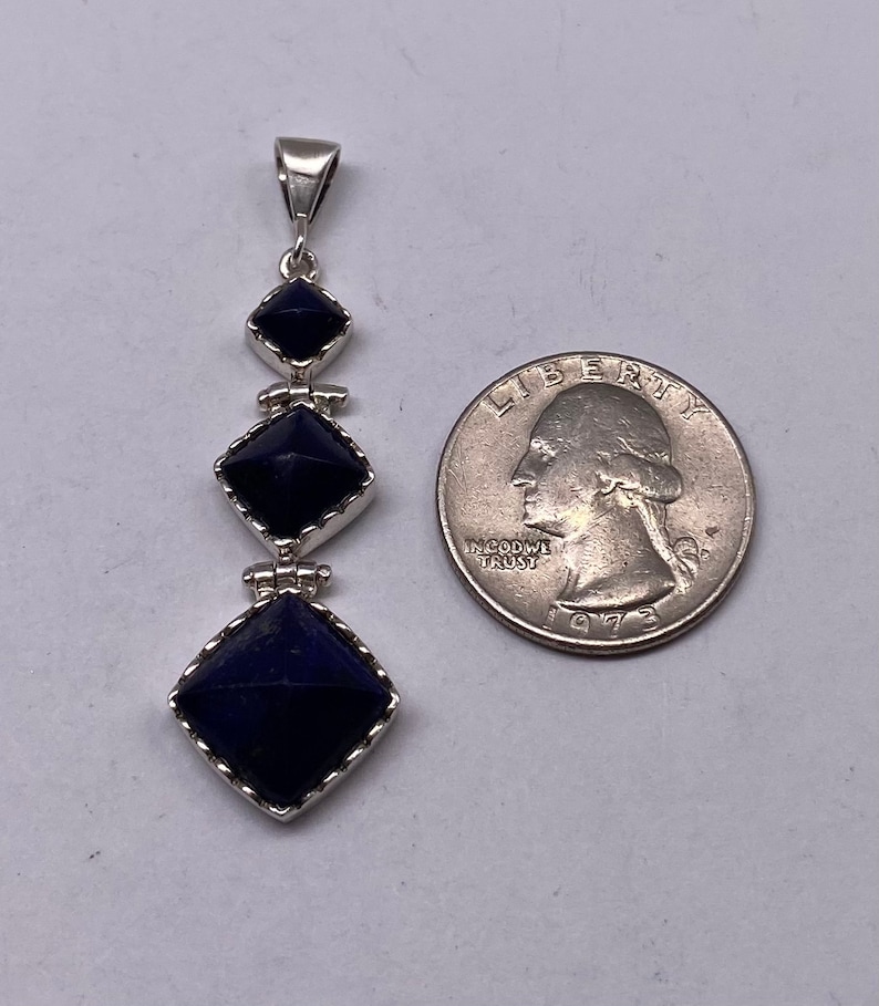 May include: A silver pendant with three dark blue lapis lazuli stones in a square shape, each stone is set in a silver frame with a scalloped edge. The pendant is hanging from a silver bail. The pendant is about the size of a US quarter.