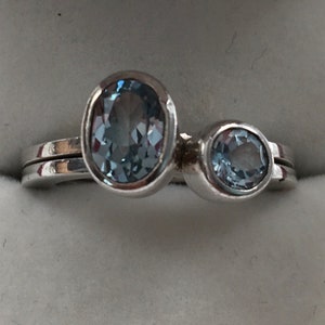 May include: A silver ring with two oval-shaped blue gemstones. The larger stone is set on the top of the ring and the smaller stone is set on the side.