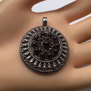 May include: A silver pendant with a circular design featuring a central cluster of dark red gemstones surrounded by intricate silverwork.