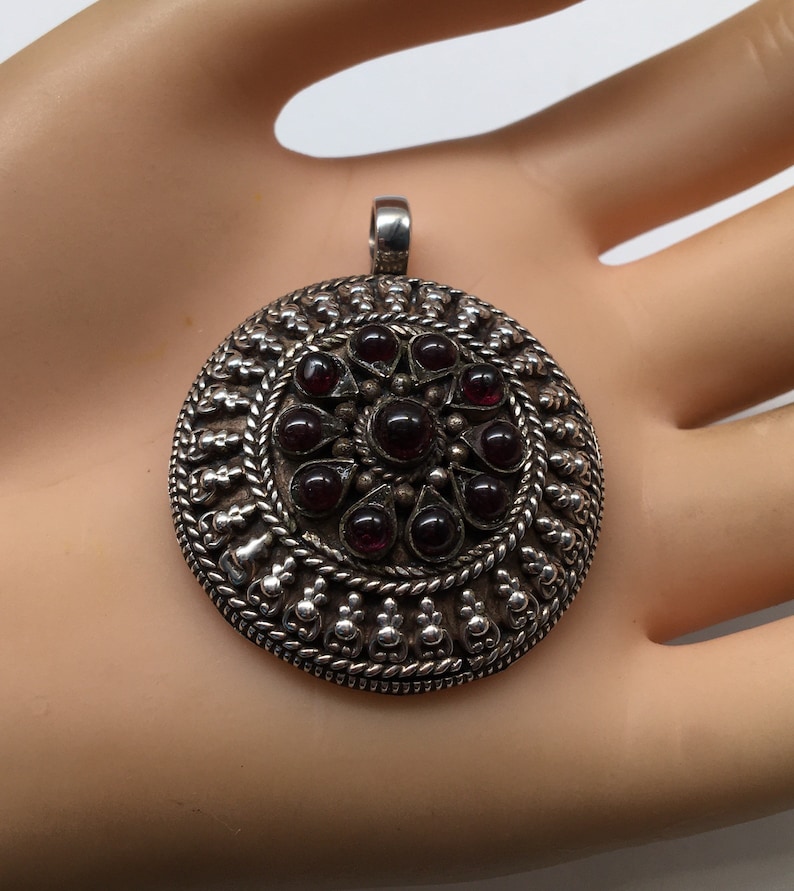 May include: A silver pendant with a circular design featuring a central cluster of dark red gemstones surrounded by intricate silverwork.