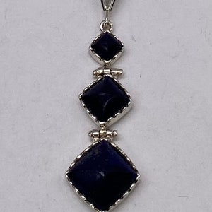May include: A silver pendant with three dark blue square-shaped stones. The stones are set in silver frames with a decorative border. The pendant is attached to a silver bail.