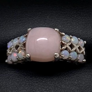 K&ouml;nnte beinhalten: Ein silberner Ring mit einem gro&szlig;en rosafarbenen Opal-Edelstein, umgeben von kleineren wei&szlig;en Opalen.