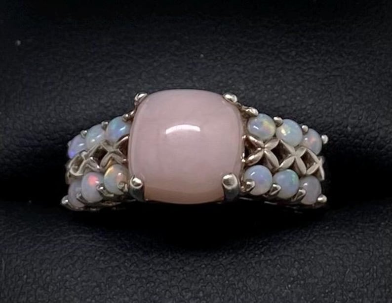K&ouml;nnte beinhalten: Ein silberner Ring mit einem gro&szlig;en rosafarbenen Opal-Edelstein, umgeben von kleineren wei&szlig;en Opalen.