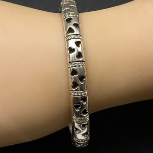 May include: A silver bangle bracelet with a heart design and small clear stones. The bracelet is on a person's wrist.