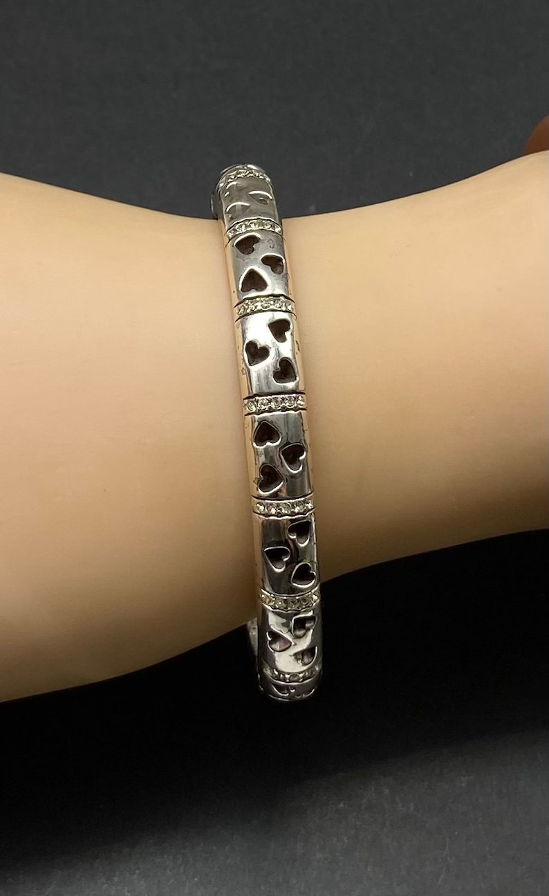 May include: A silver bangle bracelet with a heart design and small clear stones. The bracelet is on a person's wrist.