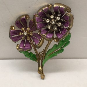 Puede incluir: Un broche de tono dorado con dos flores de esmalte morado con centros de pedrería y hojas de esmalte verde.