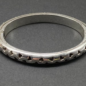 May include: A silver bangle bracelet with a hinged closure and a design of small heart cutouts around the entire circumference.