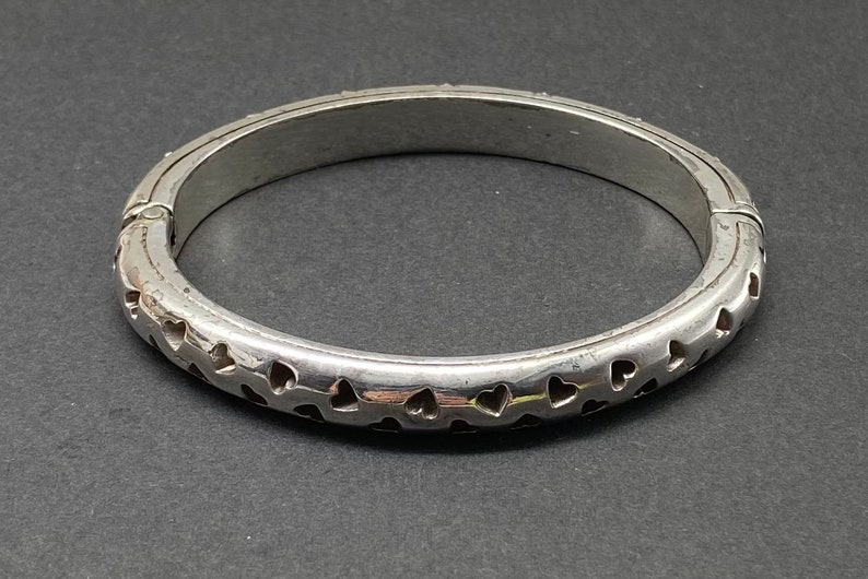 May include: A silver bangle bracelet with a hinged closure and a design of small heart cutouts around the entire circumference.