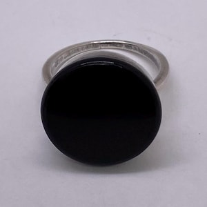 Op de afbeelding: Een zilveren ring met een grote, ronde, gepolijste zwarte steen. De ringband is dun en metaalachtig, wat contrasteert met het gladde, donkere oppervlak van de steen. De ring staat tegen een effen, lichte achtergrond.
