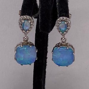 May include: A pair of silver-toned drop earrings. Each earring features a small, oval-shaped blue gemstone at the top, surrounded by small clear stones. A larger, square-shaped blue gemstone hangs below. The gemstones have a shimmering, iridescent quality.