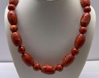 May include: A coral-colored necklace displayed on a white bust. The necklace features a series of oval and round beads in varying sizes. The beads have a natural, textured appearance, and the color is a warm, reddish-orange.