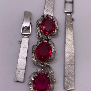 May include: A silver-toned metal bracelet featuring three large, oval-shaped, faceted red stones. Each stone is surrounded by a textured, leaf-like metal setting. The bracelet includes two separate, matching silver-toned metal strap segments.