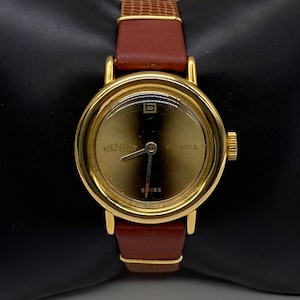 Puede incluir: Un reloj de pulsera dorado con una correa de cuero marrón. La esfera del reloj es dorada y tiene agujas negras y marcas de números romanos. La esfera del reloj también dice "MENTOS 17 JEWELS SWISS".