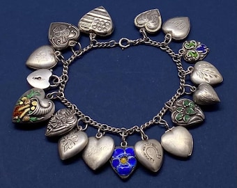 May include: A silver charm bracelet with heart-shaped charms. The charms include engraved designs, a lock, a horseshoe, and a blue flower with a yellow center. Some charms have enamel details in green, red, and blue. The bracelet is on a dark blue background.