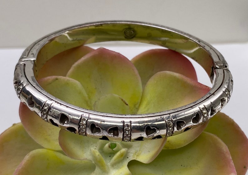 May include: A silver bangle bracelet with a heart design and clear stones. The bracelet is hinged and has a polished finish.