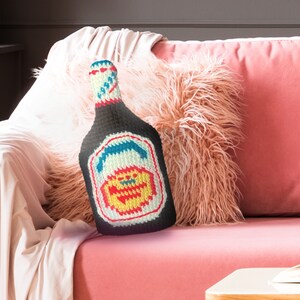 May include: A crocheted bottle-shaped cushion with a colourful label. The cushion is sitting on a pink sofa with a fluffy white throw blanket.