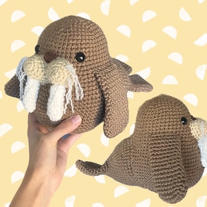 May include: Two crocheted walrus plushies, one brown with white tusks and a beige nose, the other brown with a beige nose. The walrus on the left is being held by a hand.