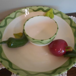 May include: A white ceramic serving platter with a green wavy border and a small matching bowl in the center. The platter features a green chili pepper and a red radish as decorative accents. A charming addition to any kitchen.