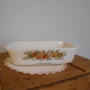 May include: A rectangular, white casserole dish with a colorful vegetable design and the text "LE BON POTAGER". The dish has handles and is set on a white, lace doily. The dish is on a wooden surface.