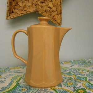 May include: A vintage, light orange ceramic coffee pot with a lid, handle, and spout. The pot has a fluted design and sits on a floral patterned tablecloth. A woven wall hanging is in the background.