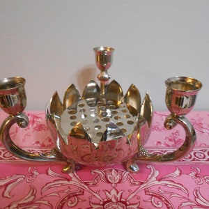 May include: A silver-colored centerpiece with a floral design, featuring two candle holders and a central flower arranger. The piece has a reflective surface and is set on a pink floral patterned surface.