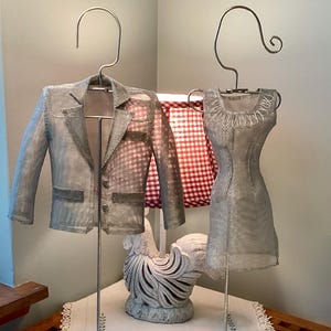 May include: Two miniature clothing forms made of silver-colored wire mesh, one shaped like a blazer with lapels and the other like a sleeveless dress with a round neckline. Each form hangs from a silver metal hook on a thin vertical stand.