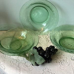 May include: Four light green glass bowls and plates with etched spiral designs. The bowls and plates are arranged on a white lace tablecloth, with a bunch of black grapes in the foreground. The glassware is vintage.