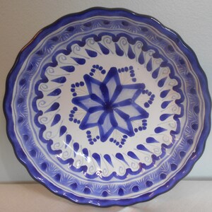 May include: A white ceramic plate with a blue floral design. The plate has a scalloped edge with a dark blue border. The center features a blue starburst design. The plate is suitable for serving food.
