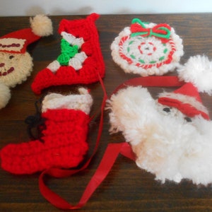 May include: A collection of handmade Christmas ornaments. The ornaments include a Santa face, a Santa hat, two red stockings, and a wreath. The ornaments are made of crochet and have red, white, green, and black accents.
