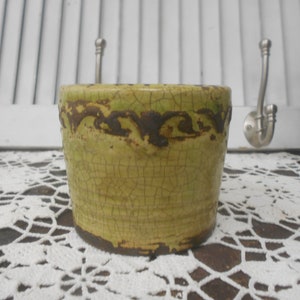 May include: A small, cylindrical ceramic pot with a textured, crackled surface. The pot is primarily a pale green color, with a dark brown band around the top and bottom. The design has a rustic, aged appearance, suggesting it could be used as a decorative planter.
