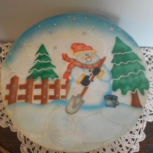 May include: Round, hand-painted winter scene featuring a snowman with a red hat and scarf, holding a shovel. The design includes green pine trees, a brown fence, and a blue bucket. The background is a gradient of blue and white, with snowflakes.