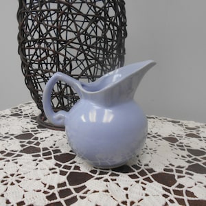 May include: A small, light blue ceramic pitcher with a curved handle and spout. The pitcher sits on a white lace doily, with a dark brown woven basket in the background. The pitcher is a soft, pastel color.