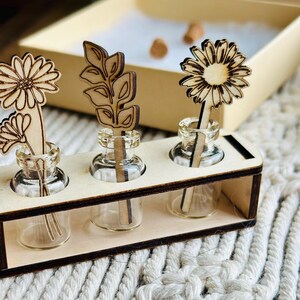 May include: Wooden stand with three small glass bottles, each containing a wooden cutout of a flower or leaf. The stand is made of light brown wood and has a natural finish.