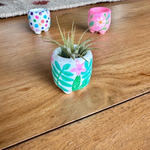 May include: Three small, hand-painted ceramic pots with a whimsical design. One pot is white with colorful polka dots, another is pink with floral accents, and the third is white with pink flowers and green leaves. The pots are on a wooden surface.