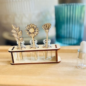 May include: A wooden stand holding three small clear glass vials, each containing a laser-cut wooden plant topper shaped like a leaf, a daisy, and a flower. A separate small clear glass vial with a white dropper cap sits to the right.