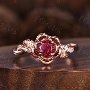 May include: Rose gold ring featuring a central, round, deep red gemstone. The ring's design incorporates a rose-shaped setting around the gemstone, with delicate leaf and small stone accents on the band. The ring is displayed on a wooden surface.