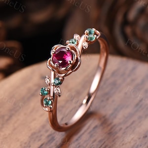 Puede incluir: Un anillo de oro rosa con una piedra preciosa roja de corte redondo en el centro, engastada en una montura de metal con forma de flor. La banda está adornada con pequeñas piedras verdes y motivos de hojas esculpidas a los lados.