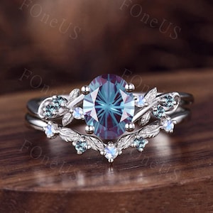 May include: A silver ring set featuring an oval-cut, color-changing gemstone. The ring is accented with small, round gemstones and leaf-like details. The band is split, creating a unique design. The ring is set on a wooden surface.