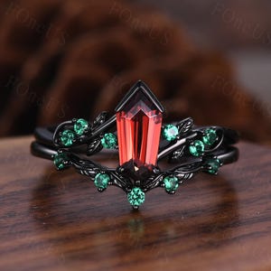 Könnte beinhalten: Schwarzes Ring-Set mit einem markanten, geometrisch geschliffenen, roten Edelstein. Der Ring ist mit kleinen, runden, grünen Edelsteinen und Blattmotiven verziert. Das Band weist ein aufwendiges, von der Natur inspiriertes Design auf.