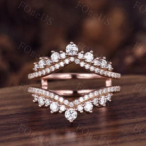 May include: Rose gold ring guard with a crown design, encrusted with numerous small, clear gemstones. The ring guard has a V-shaped design, with a larger gemstone at the center of each V.