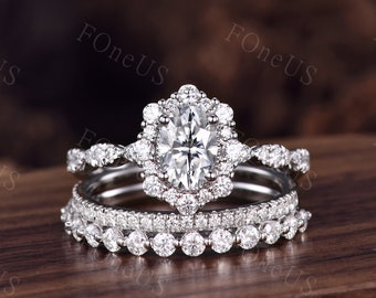 May include: A three-piece bridal ring set featuring a large oval-shaped center stone surrounded by a halo of smaller stones. The band is a delicate, twisted design with smaller stones set along the band.