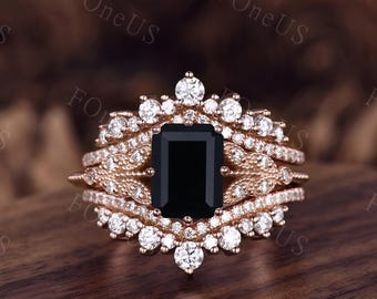 May include: A rose gold engagement ring set with a large black emerald cut gemstone surrounded by smaller clear gemstones. The ring has a delicate, intricate design with multiple bands.