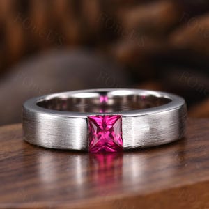 May include: A silver ring with a brushed finish, featuring a square, vibrant pink gemstone. The ring is set on a wooden surface, showcasing the contrast between the metal and the gemstone.