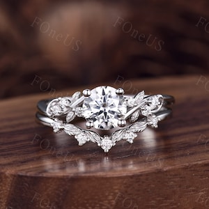 May include: A silver engagement ring set with a large, round, clear gemstone. The band features a floral design with smaller accent stones. The ring sits on a wooden surface.