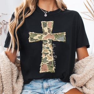 May include: Black t-shirt featuring a camouflage cross design with frayed edges. The cross is made of patches in shades of green, brown, and tan. The words "He is Risen" are written vertically on the right side of the cross in a script font.